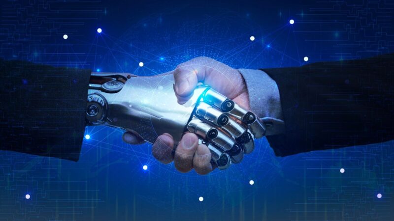 Emberi és robotkéz fog kezet, ami az ember és a mesterséges intelligencia együttműködését szimbolizálja kék digitális háttér előtt.