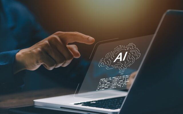 Egy kéz mutat egy laptop képernyőjén megjelenő mesterséges intelligencia (AI) ikonra, ami egy agyat ábrázol áramköri mintázattal.