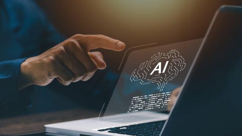 Egy kéz mutat egy laptop képernyőjén megjelenő mesterséges intelligencia (AI) ikonra, ami egy agyat ábrázol áramköri mintázattal.