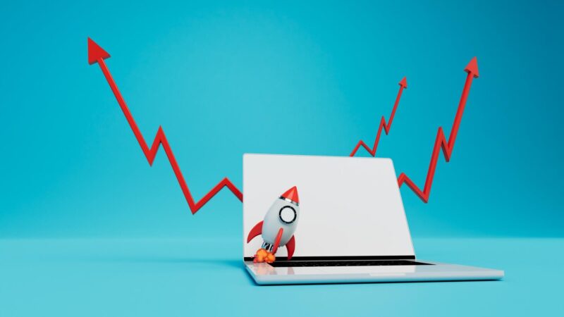 Egy laptop képernyőjén egy felszálló rakéta látható, mögötte piros, felfelé ívelő nyíl alakú grafikon. Kék háttér. SEO és rangsorolás növekedés.