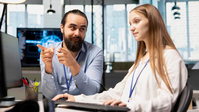 Két munkatárs, egy férfi és egy nő, számítógép előtt ülve beszélgetnek egy irodában, valószínűleg mesterséges intelligencia témában.
