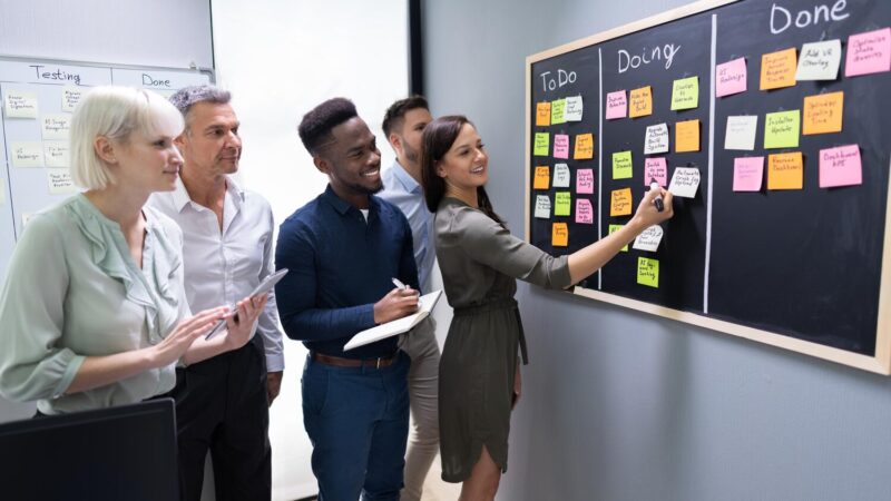 Kollégák egy kanban-táblán rendezik a feladatokat öntapadós cetlikkel