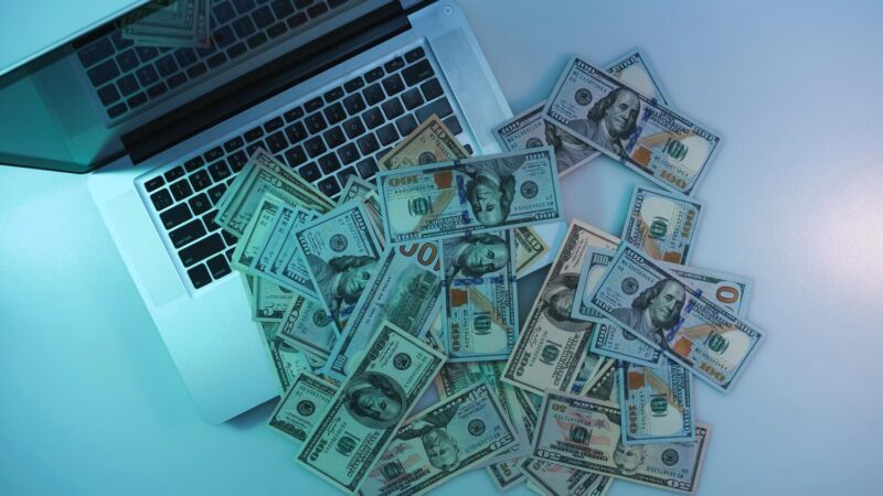 Nyitott laptop billentyűzetén és mellette szétszórt százdolláros bankjegyek felülnézetből.