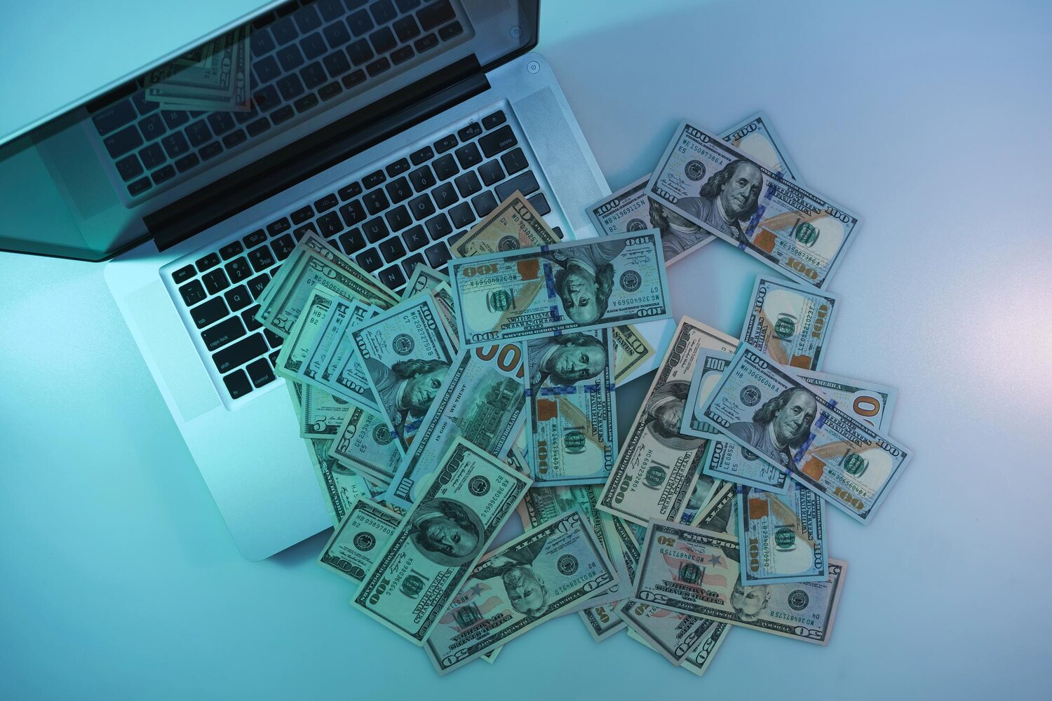 Nyitott laptop billentyűzetén és mellette szétszórt százdolláros bankjegyek felülnézetből.