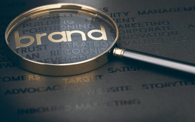 Nagyító fókuszál az arany színű brand feliratra és kapcsolódó marketing kulcsszavakra fekete háttéren.
