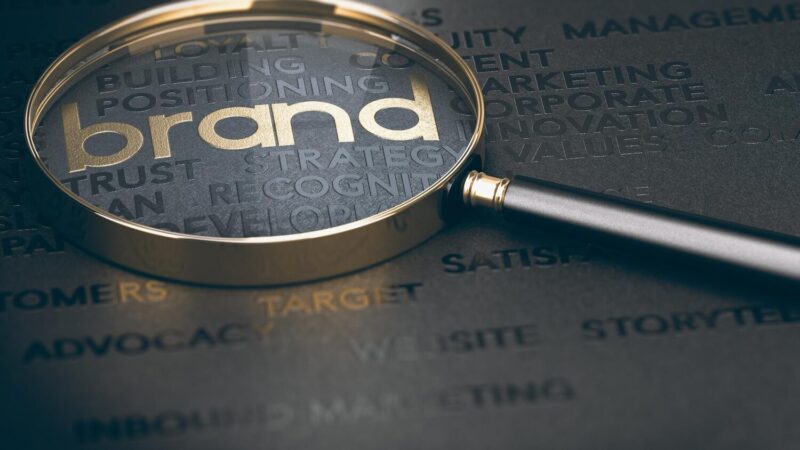Nagyító fókuszál az arany színű brand feliratra és kapcsolódó marketing kulcsszavakra fekete háttéren.