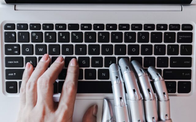 Emberi kéz és robotszerű fém kézfej gépel egymás mellett egy fehér laptop billentyűzetén felülnézetből.