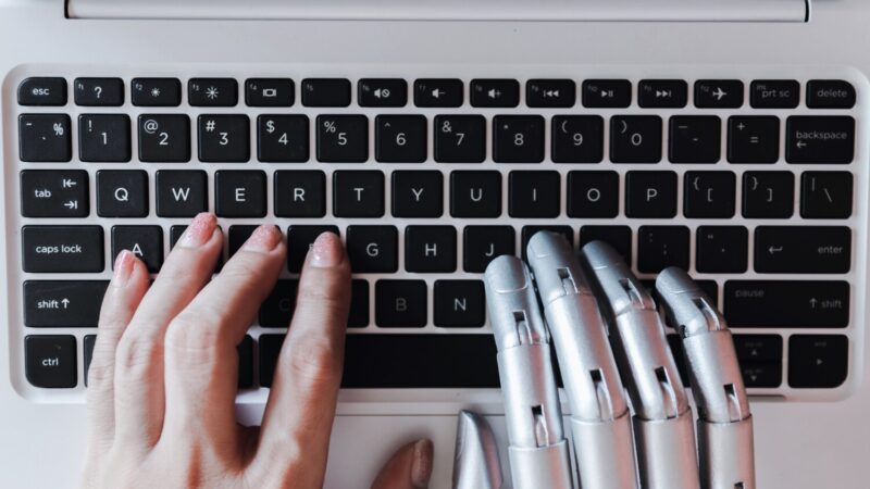 Emberi kéz és robotszerű fém kézfej gépel egymás mellett egy fehér laptop billentyűzetén felülnézetből.