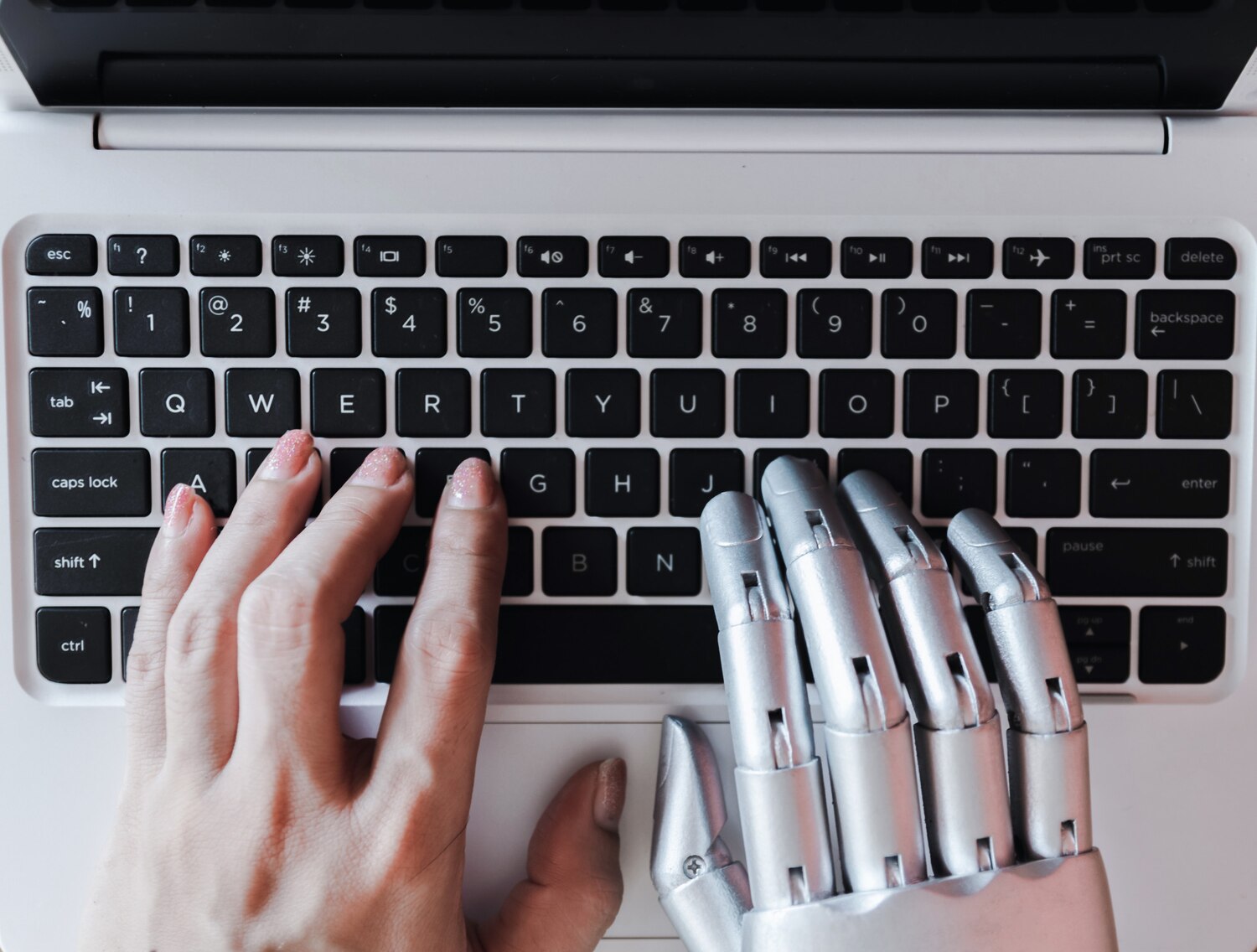 Emberi kéz és robotszerű fém kézfej gépel egymás mellett egy fehér laptop billentyűzetén felülnézetből.