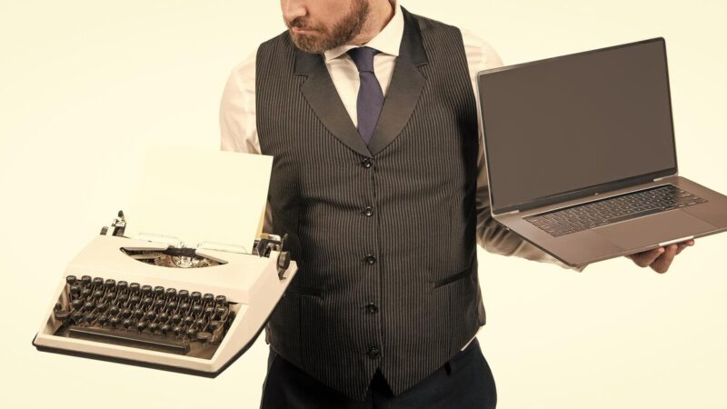 Férfi az egyik kezében mechanikus írógépet, a másikban modern hordozható laptopot tart.